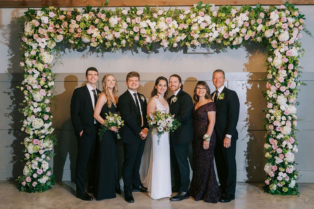 A groom and bride with their family members, smiling in front of a floral background display at a garden wedding venue named Fleetwood Farm Winery in Leesburg, Virginia.