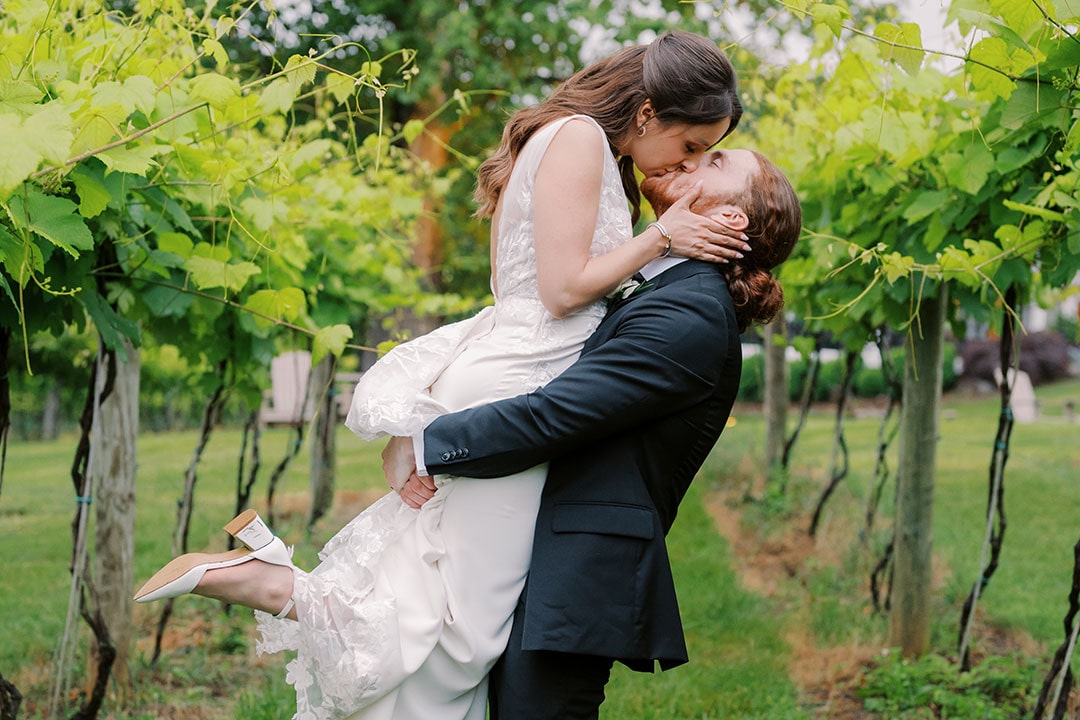 A groom lifting up a bride and kissing her amidst vineyards at a winery wedding vineyard in Leesburg, Virginia.