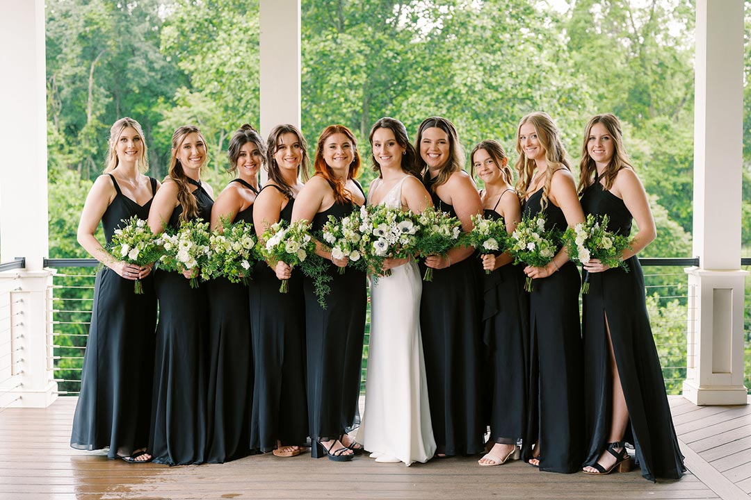 A bride in a white wedding dress, surrounded by her bridesmaids, all dressed in black and holding flowers on an upper terrace area at a Virginia winery wedding venue.