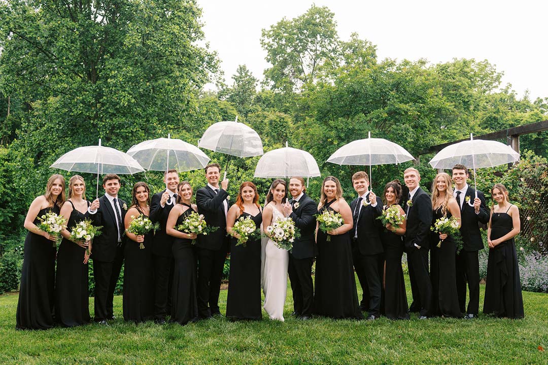 A groom and bride smiling, surrounded by their wedding party, all dressed in black, holding umbrellas at a winery wedding venue in Leesburg, Virginia.