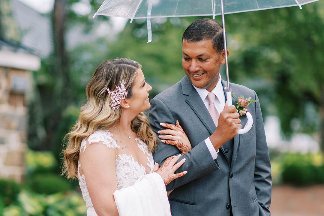 A man in a suit holding an umbrella while locked in arms with a smiling bride at a garden wedding venue in Leesburg, Virginia.