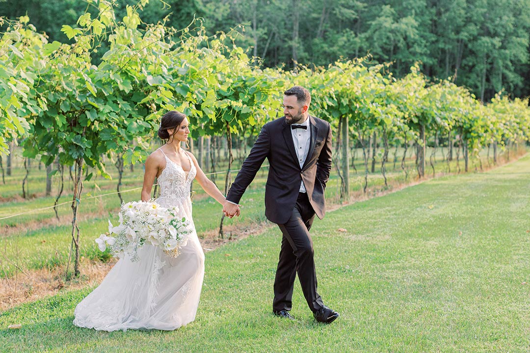 A bride and groom holding hands, walking through the vineyards at Fleetwood Farm Winery, a winery wedding venue located in Leesburg, Virginia.