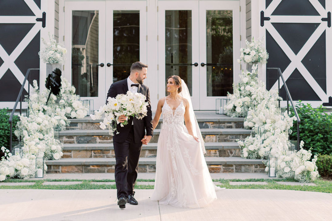 A groom holding flowers and holding hands with a smiling bride outside of Fleetwood Farm Winery, a winery wedding venue in Leesburg, Virginia.