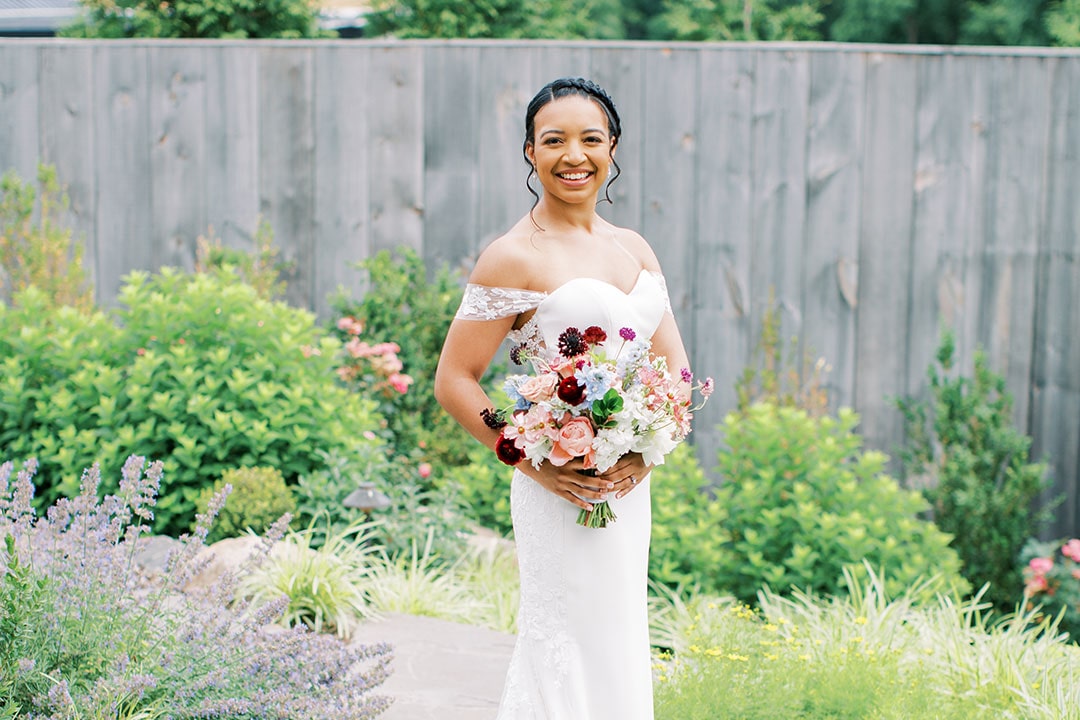 A bride in her wedding dress, smiling near a garden at Fleetwood Farm Winery, an outdoor wedding venue in the Brambleton, VA area.