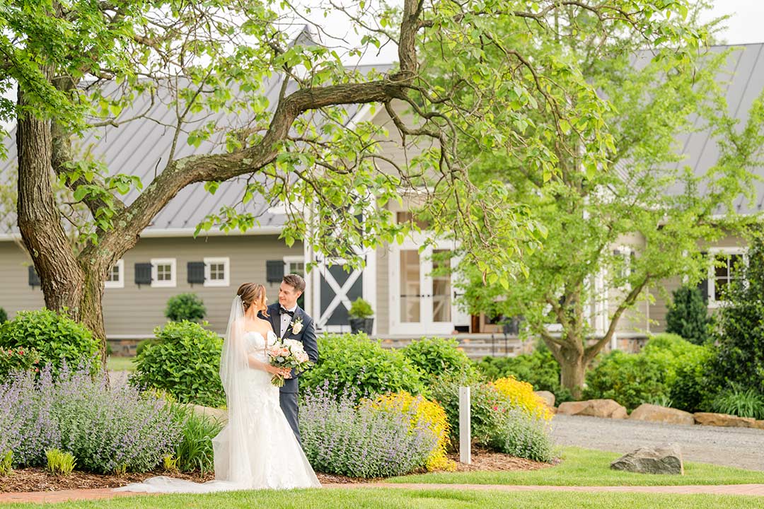 A bride and groom looking at each other, smiling, near a blooming garden area and tree at Fleetwood Farm Winery, a Leesburg wedding venue & winery.