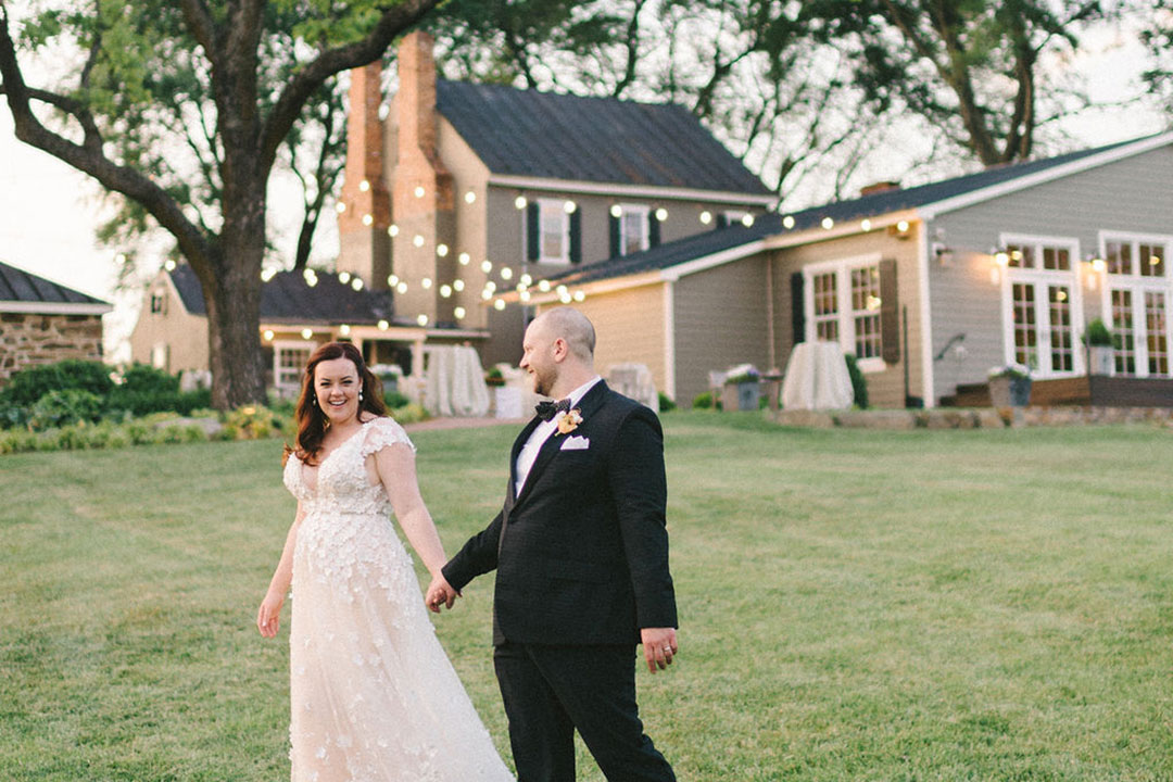 A groom holding hands with his bride in a grassy area outside of Fleetwood Farm Winery, an outdoor wedding venue in Northern Virginia.