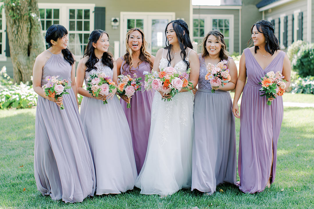 Six bridesmaids in dresses, holding bouquets, smiling at one another in a grassy area at Fleetwood Farm Winery, a winery wedding venue in Loudoun County, VA.