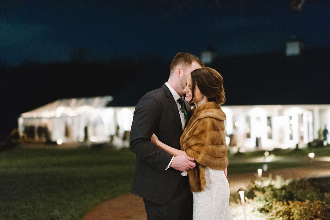 A bride smiling, hugging her groom outside along a brick walkway at Fleetwood Farm Winery, an outdoor wedding venue in Loudoun County, Virginia.