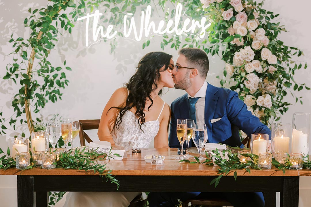 A bride and groom seated, kissing at a 