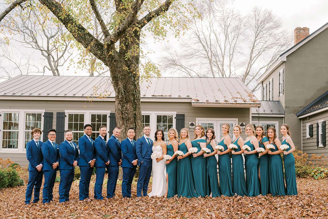 Groomsmen and bridesmaids dressed in blue suits and dresses, lined up for a picture amidst fall foliage outside of Fleetwood Farm Winery in Leesburg, Virginia.