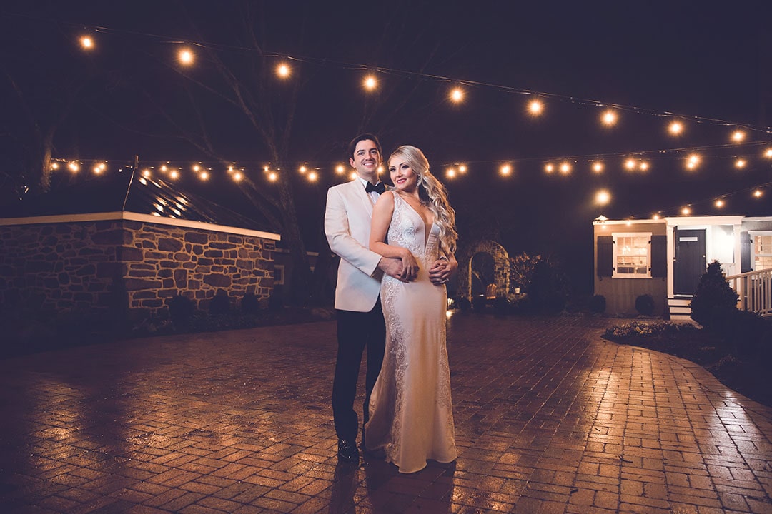 A smiling bride & groom posing together with bulb lights overhead at a winter wedding venue in Leesburg, Virginia.