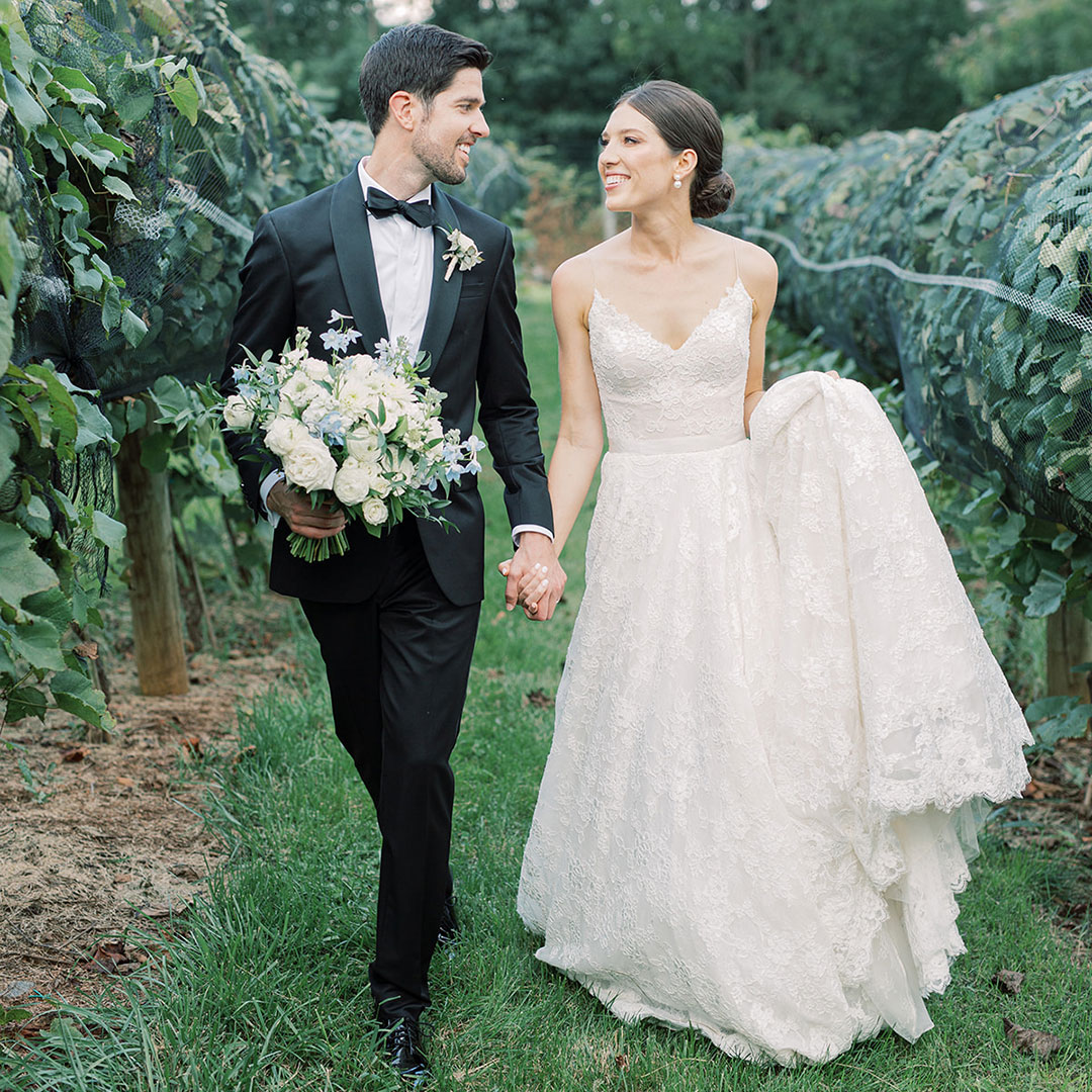 A groom and bride holding hands, smiling at each other as they walk through the vineyards at a top Virginia winery called Fleetwood Farm Winery.